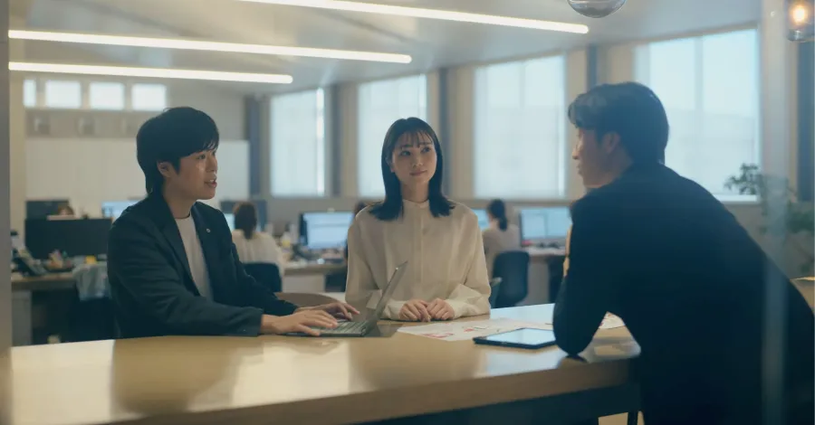 写真：ミーティングテーブルで、PCやタブレットを広げて会議をしている3名の社員